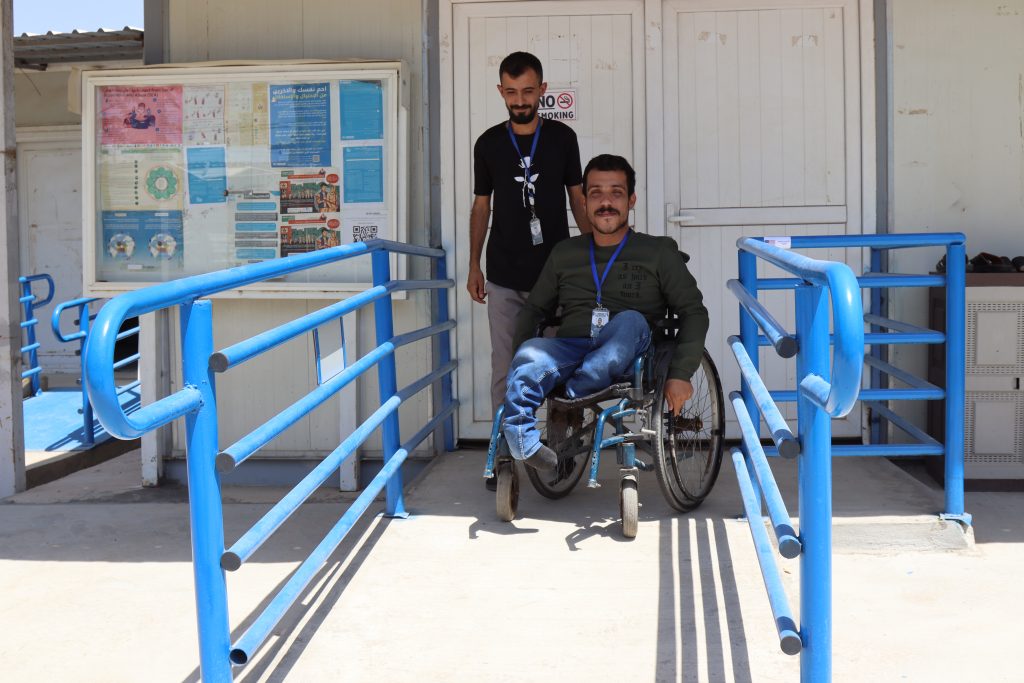 Two men smile at the top of a concrete wheelchair ramp with blue railings. One is sitting in a wheelchair while the other is standing