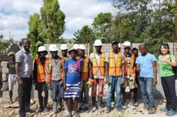 Group of Peace Winds Haitian staff and volunteers pose in front of a construction site wearing reflective vests and white hard hats