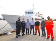 Seven Kenshin ship staff members pose on a dock in front of the Kenshin, a large white ship