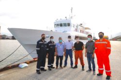 Seven Kenshin ship staff members pose on a dock in front of the Kenshin, a large white ship