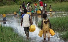 A grassland in Mozambique is flooded, and a group of people wade through the water carrying yellow jugs