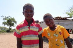 Two Mozambican boys, young teenagers, smile in the foreground. One is wearing a yellow shirt and one is wearing a red striped shirt. They are standing outside in a background of trees and a dirt ground.