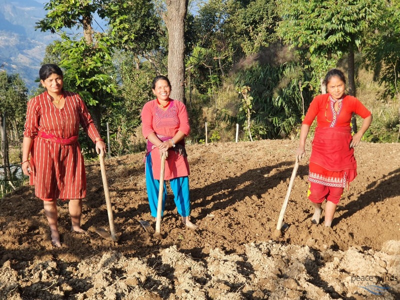 Three Nepalese women wearing red clothing stand in a dirt field holding garden tools