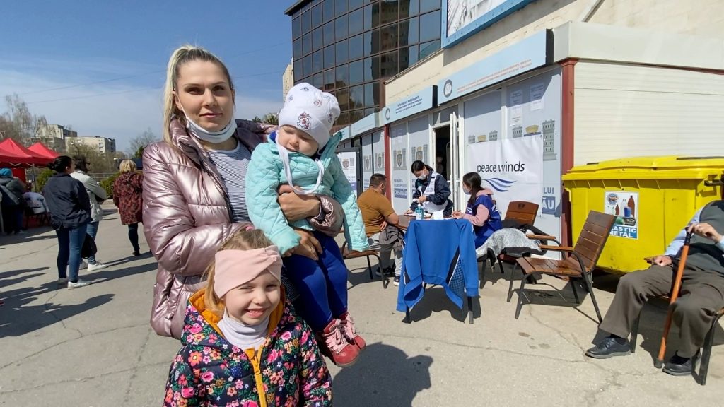 Ukrainian woman wearing a winter coat holds a baby also wearing a winter coat and hat with a young, smiling girl of about 6 standing in front of her. "Peace Winds" branded distribution tables are set up in the background