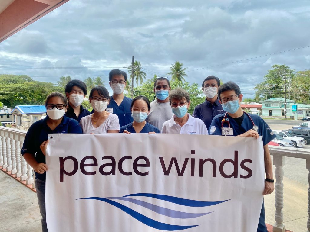 Nine Palauan and Japanese Peace Winds staff members stand in a group for a photo wearing masks an holding a large banner with the Peace Winds logo