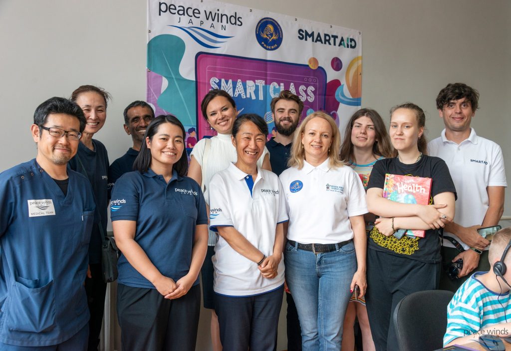 11 staff members from Peace Winds and partner organizations Phoenix and Smart Aid pose for a group photo in front of a colorful "Smart Class" banner