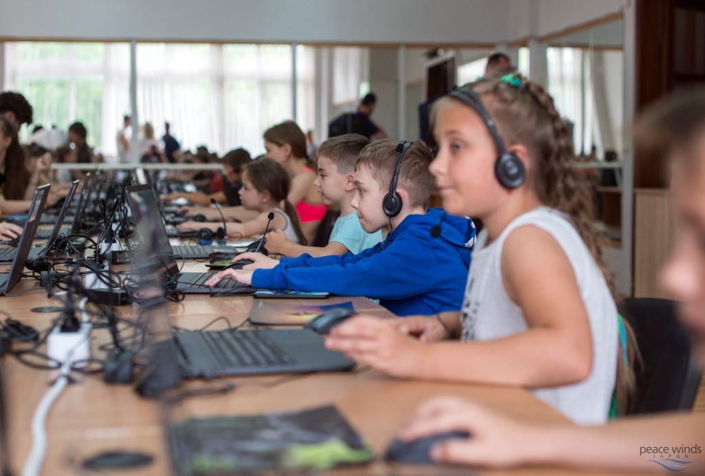 Several dozen Ukrainian children sit at a long table using headphones and laptop computers