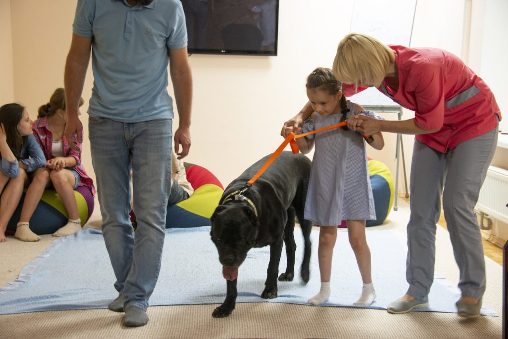 A young girl holds onto the orange leash of a black lab with help from a woman wearing pink scrubs