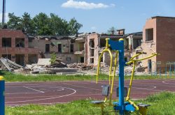 A school building stands with half of its walls destroyed, rubble sitting next to the building with a basketball court and playground equipment in the foreground