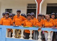 A large group of Haitian women wearing orange shirts and dark blue baseball caps stands together on a white and blue balcony