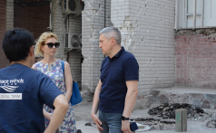 Woman wearing a floral dress and sunglasses, man in a blue polo shirt, and Peace Winds employee with his back to the viewer (wearing a Peace Winds branded t-shirt) stand in the street having a conversation near the collapsed wall of a building