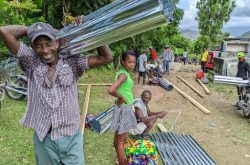 smiling Haitian man wearing a plaid shirt carries a roll of sheet metal while other Haitian men and women stand around with wood planks and sheet metal in the background