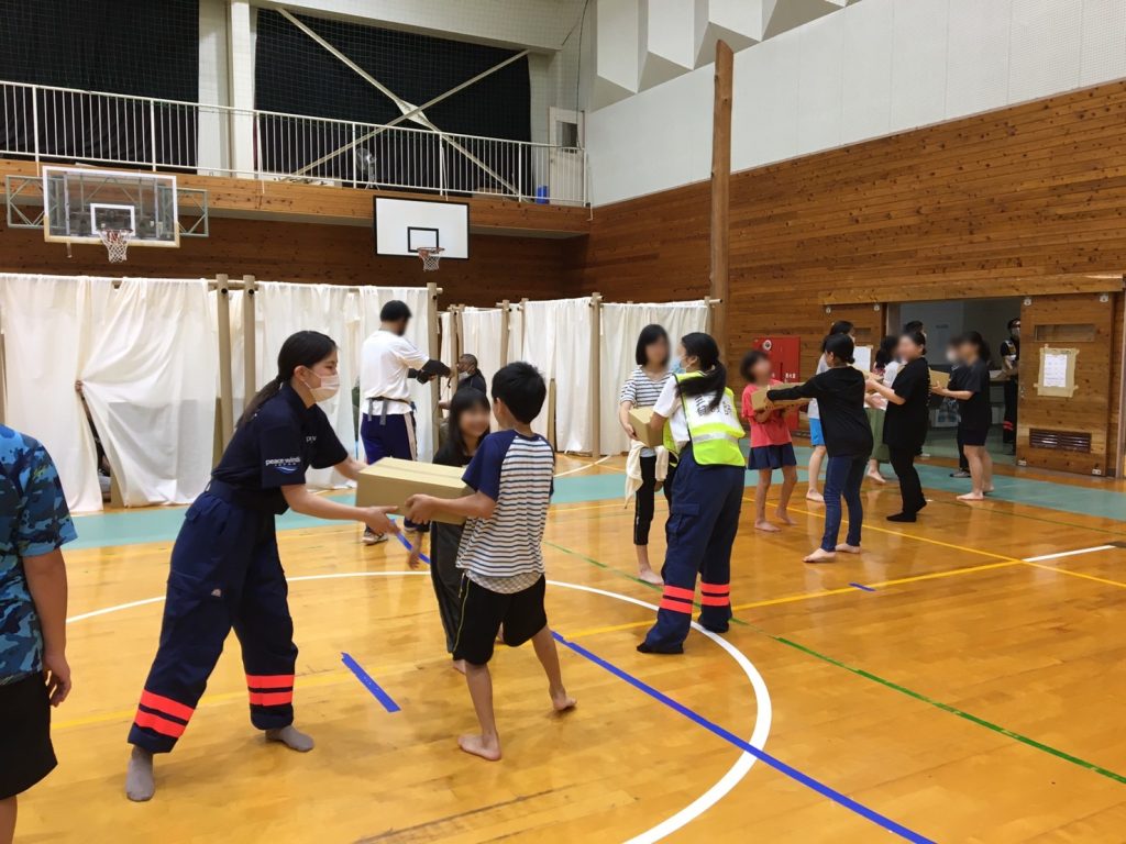 Two Japanese women wearing Peace Winds uniforms pass cardboard boxes to a line of children standing in a school gymnasium