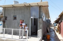 A Syrian refugee man holds a toddler and stands with an older boy in front of a gray cinderblock home with a white railing