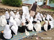 About two dozen girls wearing black clothing and white headscarves sit on a blue and yellow patterned tarp outside in the dirt
