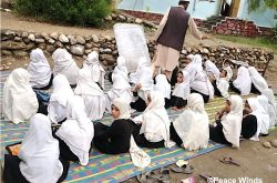 About two dozen girls wearing black clothing and white headscarves sit on a blue and yellow patterned tarp outside in the dirt