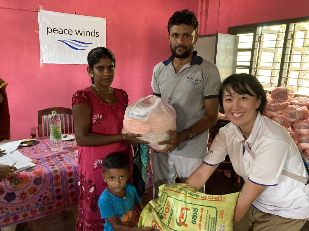 A Sri Lankan woman wearing a pink dress, a child wearing a blue t-shirt, a man wearing a gray "Peace Winds" t-shirt, and a Japanese woman wearing a white "Peace Winds" t-shirt hold bags of nonperishable foods