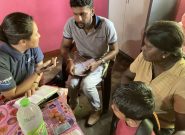 A Sri Lankan woman wearing a pink dress, a child wearing a blue t-shirt, a man wearing a gray "Peace Winds" t-shirt, and a Japanese woman wearing a white "Peace Winds" t-shirt hold bags of nonperishable foods