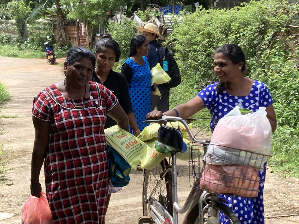 A group of Sri Lankan women walks along a path surrounded by green bushes and carries white and yellow bags of nonperishable food
