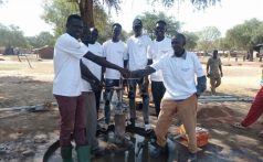 7 south sudanese men wearing white "peace winds" shirts stand around a water pump