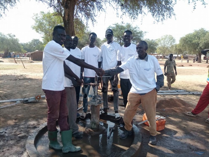 7 south sudanese men wearing white "peace winds" shirts stand around a water pump