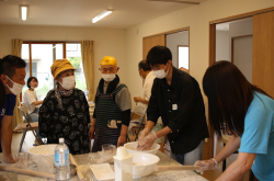 Five Japanese people wearing white face masks stand around a table with bowls, flour, and supplies for making noodles