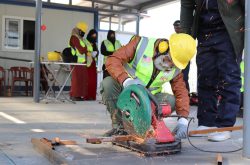 a Syrian refugee man with a white beard, yellow hardhat, neon safety vest, orange long sleeve shirt, and green pants crouches down to use a circular saw that sprays orange sparks