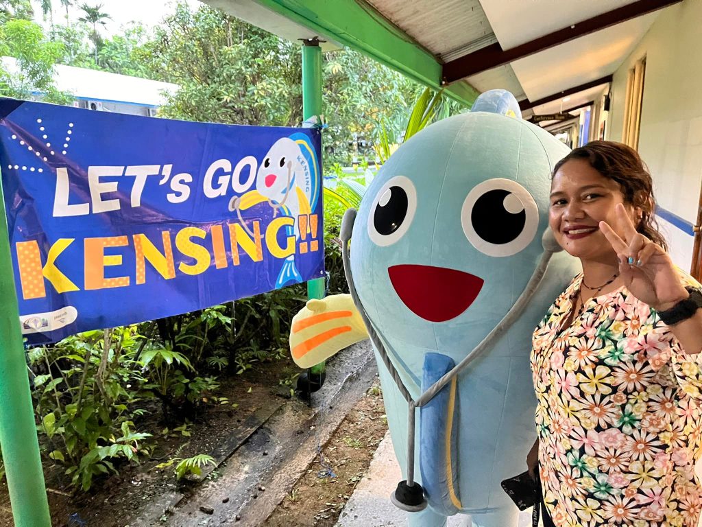 Woman wearing a flower scrubs shirt holds up a peace sign while standing with a "real-life" version of the Peace Winds Palau mascot, a person in a blue and yellow fish costume