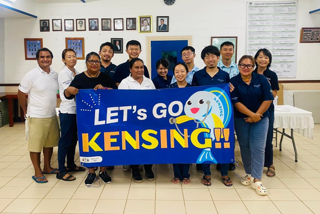 About a dozen Japanese and Palauan Peace Winds staff members wearing Peace Winds tee shirts pose with a blue banner that says "let's go kensing!!" and has a cartoon picture of a blue and yellow fish wearing a stethoscope 