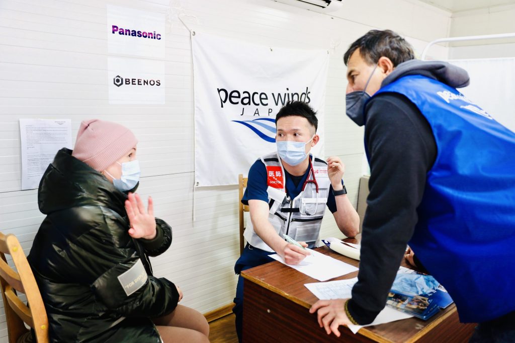 Two Peace Winds doctors speak to a middle aged Ukrainian woman around a wooden desk in a white medical tent