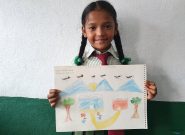 a young Nepalese girl with two braids smiles while holding the drawing she made. The draqing has blue mountains, a yellow sun, a flock of black birds, two trees, and two buildings labeled "my house" and "my kitchen."