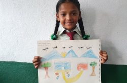 a young Nepalese girl with two braids smiles while holding the drawing she made. The draqing has blue mountains, a yellow sun, a flock of black birds, two trees, and two buildings labeled "my house" and "my kitchen."