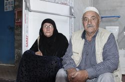 Two elderly Syrian refugees, a man and a woman, sit in front of a newly renovated bathroom. The woman is wearing a black dress and hijab, and the man is wearing a blue shirt with a white vest and white hat