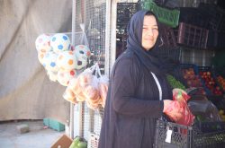 A smiling Syrian refugee woman wears a black hijab and black sweater while holding a basket at an outdoor market with fruits and vegetables. In the background on the left, there is a bag of colorful soccer balls