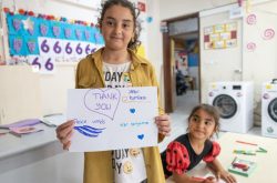 a turkish girl of about 11 smiles holding a hand-drawn "thank you" card with the peace winds and japan platform logos