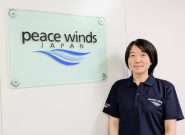 Japanese woman smiles slightly while wearing a dark blue "Peace Winds" logo polo shirt and standing next to a peace winds logo on the wall