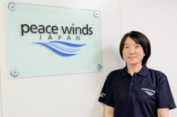 Japanese woman smiles slightly while wearing a dark blue "Peace Winds" logo polo shirt and standing next to a peace winds logo on the wall