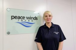 Peace Winds employee Belma Sisic wears a blue Peace Winds logo shirt and is smiling next to a "Peace Winds" logo on the wall