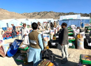 Outdoor mountainous landscape with light blue tents, piles of food packages, and several people gathered in the camp