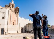 two people with "Peace Winds" logos on the back of their t-shirts stand in front of a large damaged brick mosque