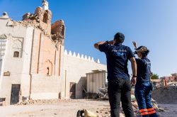 two people with "Peace Winds" logos on the back of their t-shirts stand in front of a large damaged brick mosque