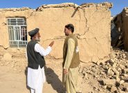 Two men talk in front of a clay/brick house with cracks and surrounded by earthquake rubble