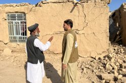 Two men talk in front of a clay/brick house with cracks and surrounded by earthquake rubble