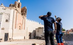 Two staff members with Peace Winds logo shirts look up at a tall, reddish-brown earthquake-damaged building
