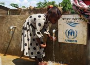 Sudanese Refugee girl wearing a gray dress with black polka dots rinses her hands under a faucet outside next to a Peace Winds and UNHCR plaque