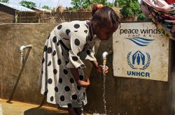 Sudanese Refugee girl wearing a gray dress with black polka dots rinses her hands under a faucet outside next to a Peace Winds and UNHCR plaque