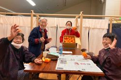 Three elderly Japanese evacuees sit at a table, smiling and waving to the camera while holding oranges. A Peace Winds staff member in a red jacket stands at the end of the table, smiling and holding a box of oranges
