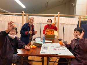 Three elderly Japanese evacuees sit at a table, smiling and waving to the camera while holding oranges. A Peace Winds staff member in a red jacket stands at the end of the table, smiling and holding a box of oranges