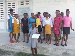Large group of Haitian women all wearing colorful clothing stands behind a smiling woman wearing a gray dress and holding a piece of white fabric
