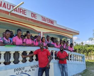 Large group of Haitian teenagers wearing pink shirts stands on the blue and white balcony of a community center behind two smiling men wearing red shirts